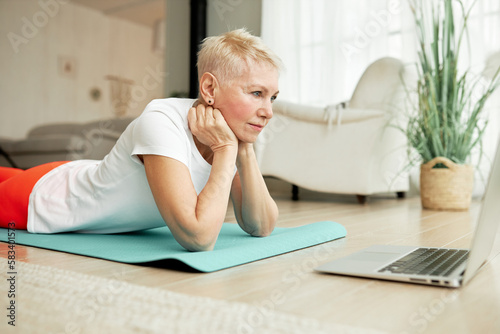 Elderly beautiful healthy slim female in ports clothes watching videos of training exercises with tips and advice and rules of proper nutrition lying on floor on mat in front of laptop