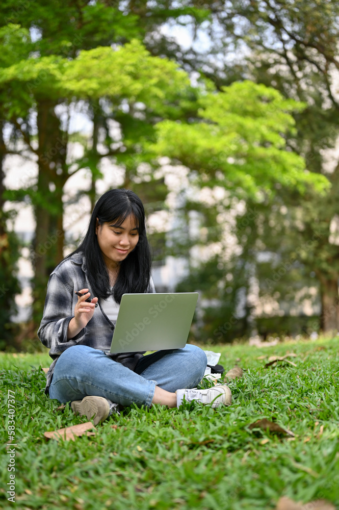 Pretty Asian female college student using her laptop and sitting on grass in the campus park. Pretty Asian female college student using her laptop and sitting on grass in the campus park.