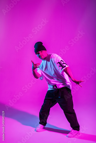 Fashion professional handsome breakdancer man with hat in stylish clothes dancing in creative studio with pink and neon lights