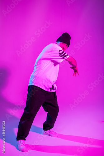 Handsome professional breakdancer man in fashion outfit with hat, white t-shirt, pants and sneakers dances in creative studio with pink and neon light