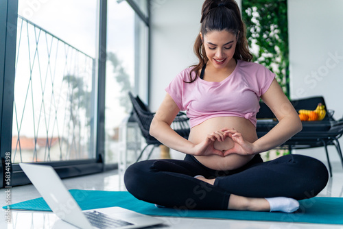 young pregnant woman siting on a yoga mat making a heart shape on her pregnant belly, excited young mother concept