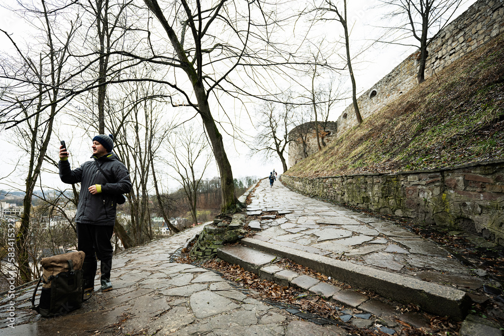 Man tourist with backpack look at phone, stand on wet path to an ancient medieval castle fortress in rain.