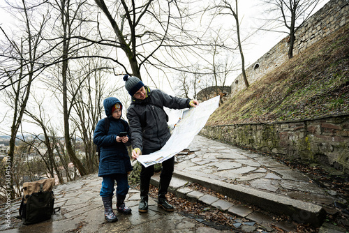 Father and son tourist look at map, stand on wet path to an ancient medieval castle fortress in rain.