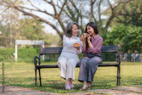 Asian mother and daughter sitting in the park happily and warmly eat food and bread together © Premreuthai