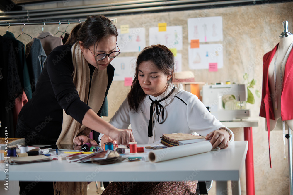 Obraz premium Asian middle-aged female fashion designer teaches a young teen trainee tailor in studio with colorful thread and sewing fabric for dress design collection ideas, professional boutique small business.