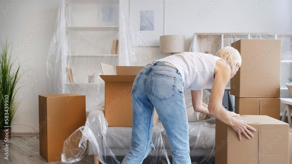 Moving day. Home relocation. Package delivery. Unrecognizable woman carrying carton boxes in light modern apartment with covered furniture. Moving day. Home relocation. Package delivery. Unrecognizable woman carrying carton boxes in light modern apartment with covered furniture.