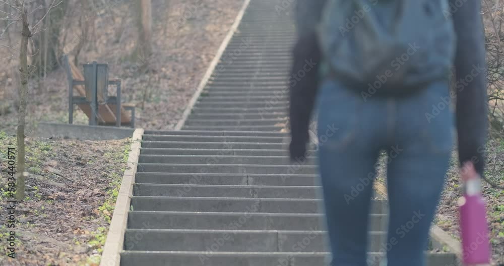 Young unrecognizable woman walks up concrete stairs. City park, daytime, springtime, back view. Flask in hand, city backpack on her back. Young unrecognizable woman walks up concrete stairs. City park, daytime, springtime, back view. Flask in hand, city backpack on her back.