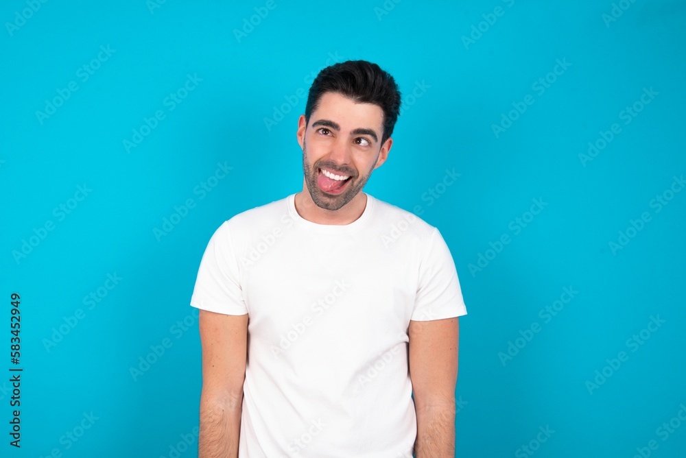 Young man wearing white T-shirt over blue studio background showing grimace face crossing eyes and showing tongue. Being funny and crazy