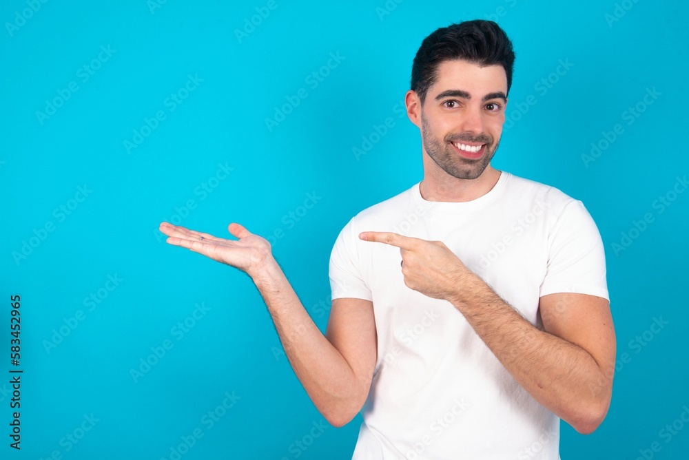 Young man wearing white T-shirt over blue studio background pointing and holding hand showing adverts Young man wearing white T-shirt over blue studio background pointing and holding hand showing adverts