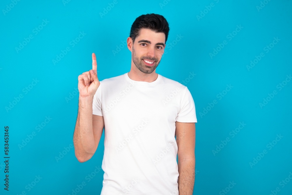 Young man standing over blue studio background showing and pointing up with finger number one while smiling confident and happy. Young man standing over blue studio background showing and pointing up with finger number one while smiling confident and happy.