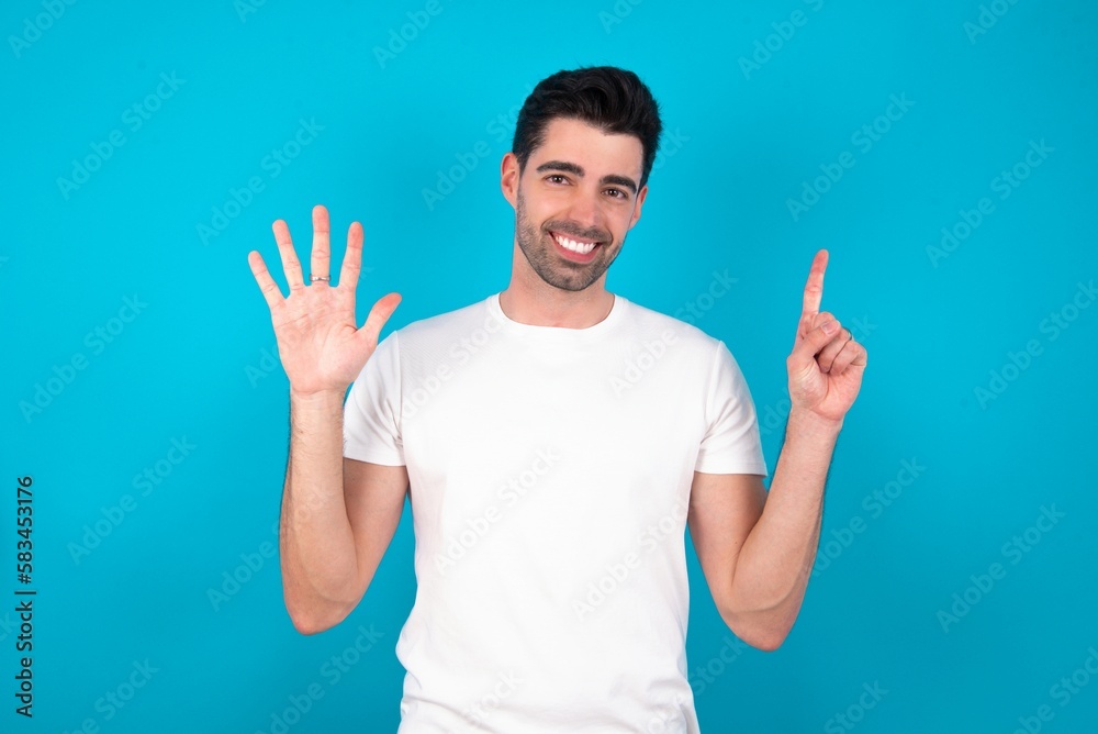 Young man standing over blue studio background showing and pointing up with fingers number six while smiling confident and happy. Young man standing over blue studio background showing and pointing up with fingers number six while smiling confident and happy.