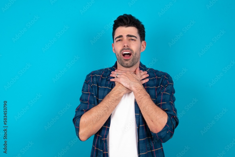 Young man standing over blue studio background shouting suffocate because painful strangle. Health problem. Asphyxiate and suicide concept. Young man standing over blue studio background shouting suffocate because painful strangle. Health problem. Asphyxiate and suicide concept.