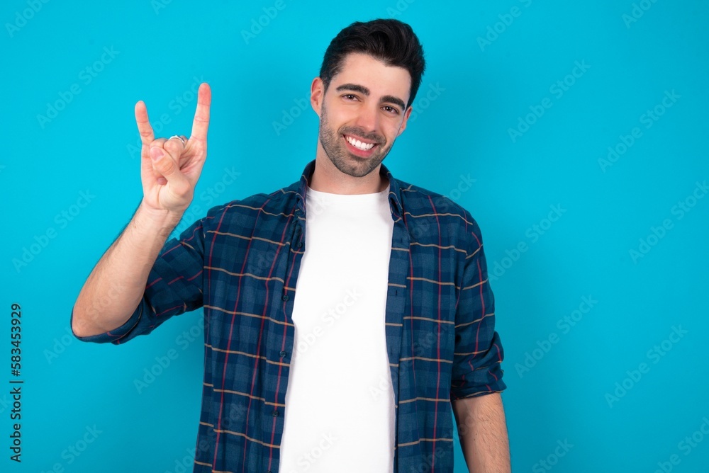 Young man standing over blue studio background doing a rock gesture and smiling to the camera. Ready to go to her favorite band concert. Young man standing over blue studio background doing a rock gesture and smiling to the camera. Ready to go to her favorite band concert.