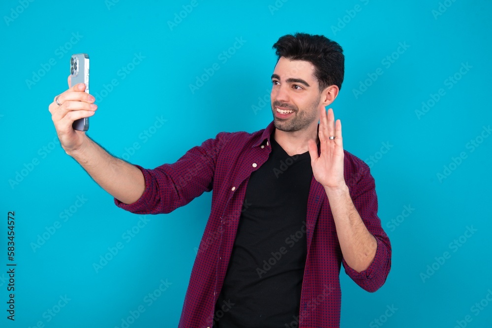 Young man standing over blue studio background holds modern mobile phone and makes video call waves palm in hello gesture. People modern technology concept Young man standing over blue studio background holds modern mobile phone and makes video call waves palm in hello gesture. People modern technology concept