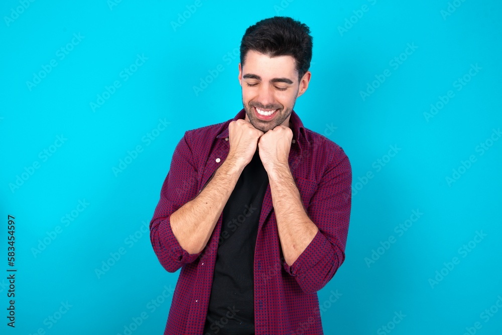 Cheerful Young man standing over blue studio background has shy satisfied expression, smiles broadly, shows white teeth, People emotions