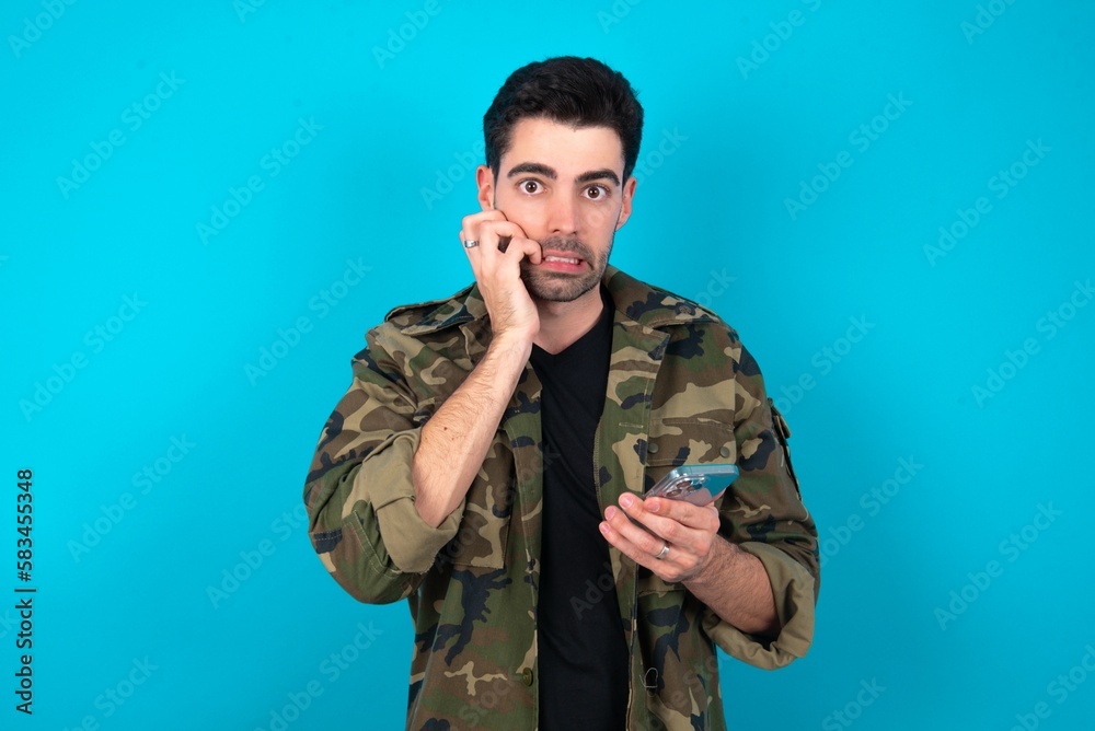 Afraid funny Young man standing over blue studio background holding telephone and bitting nails