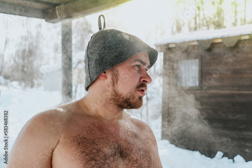The sweaty man in a hat after sauna outdoors in winter. Steam due to temperature difference. Selective focus	
