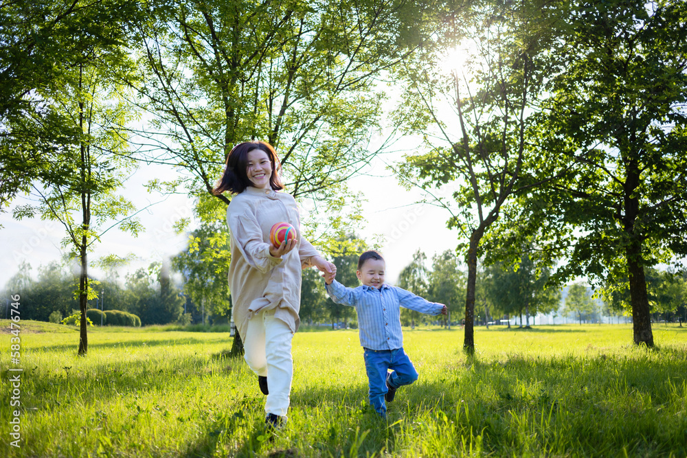 Asian mother and child play with ball, run across the field, on the grass. Kazakh family together in holiday. Happy woman and boy in summer pastime. daylight saving time