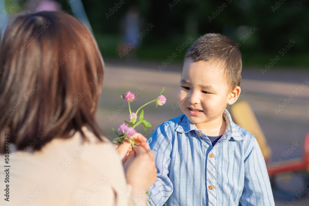 happy boy giving mom a flower. Asian kid presenting for mother bouquet of wildflowers clover. Mothers day celebration concept. Kazakh family together. Woman and child daylight saving time happy boy giving mom a flower. Asian kid presenting for mother bouquet of wildflowers clover. Mothers day celebration concept. Kazakh family together. Woman and child daylight saving time
