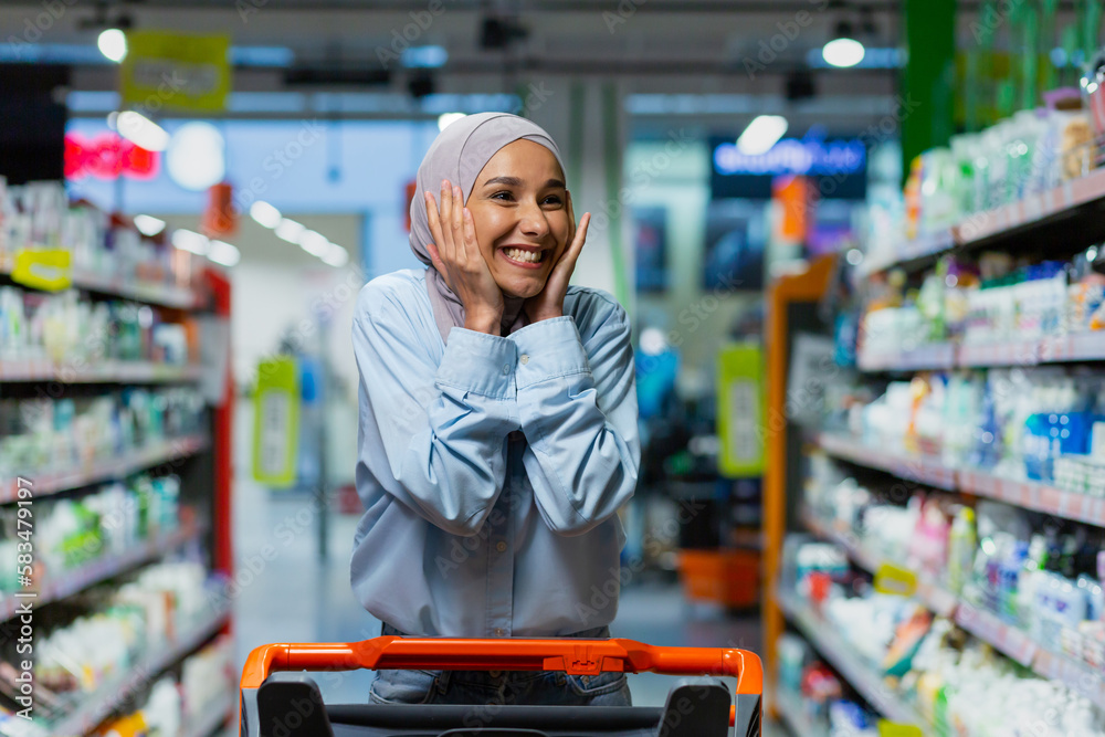 Portrait of successful satisfied woman shopper in supermarket, cheerful muslim woman in hijab with shopping trolley among shelves with goods looking at various shops.