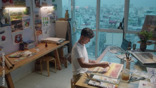 High angle wide shot of young male artist painting abstract picture with watercolors at workplace in creative studio with panoramic windows photo