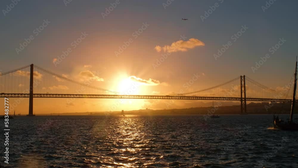 View of 25 de Abril Bridge famous tourist landmark of Lisbon connecting Lisboa and Almada on Setubal Peninsula over Tagus river with yacht silhouette on sunset and flying plane. Lisbon, Portugal View of 25 de Abril Bridge famous tourist landmark of Lisbon connecting Lisboa and Almada on Setubal Peninsula over Tagus river with yacht silhouette on sunset and flying plane. Lisbon, Portugal