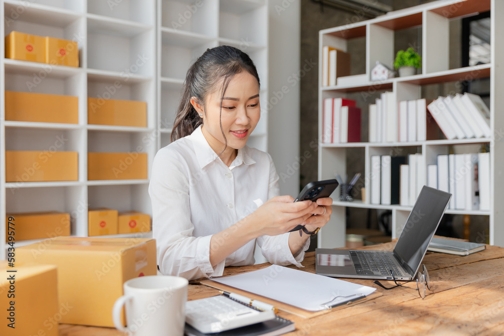 Starting an SME, an independent Asian female small business entrepreneur using a laptop and a smart phone. Cheerful success, cute smiley face. Asian woman raising her hand, packaging box, online marke