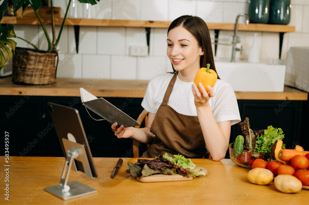 Young beautiful woman in the kitchen in an apron, fresh vegetables on the table, writes down her favorite recipes, comes up with ideas for dishes. Young beautiful woman in the kitchen in an apron, fresh vegetables on the table, writes down her favorite recipes, comes up with ideas for dishes.