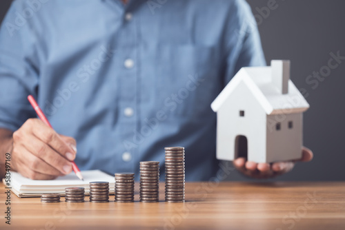 Money saving for house or Financial for real estate concept. Man making calculate note on book and coin stacked on the wooden table. Keeping fund for purchase the residence idea.