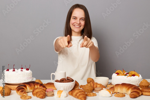 Portrait of smiling attractive woman with brown hair sitting at table isolated over gray background  pointing at camera  inviting you to have tasty unhealthy dinner  offering delicious dessert.