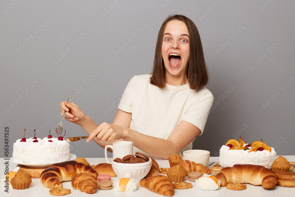 Portrait of amazed cheerful attractive woman with brown hair sitting at table isolated over gray background, eating cake, looking at camera, screaming happiness, breaking diet.