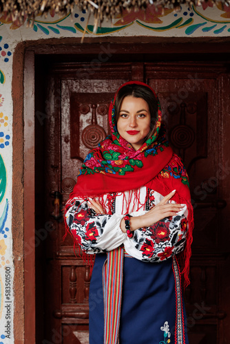 Charming smiling woman in traditional ukrainian handkerchief, necklace and embroidered dress standing at background of decorated hut. Ukraine, style, folk, ethnic culture