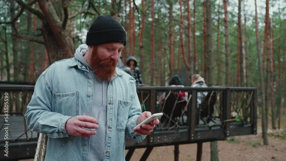 Positive bearded tourist man typing on mobile and looking at friends around the campfire in the forest Positive bearded tourist man typing on mobile and looking at friends around the campfire in the forest