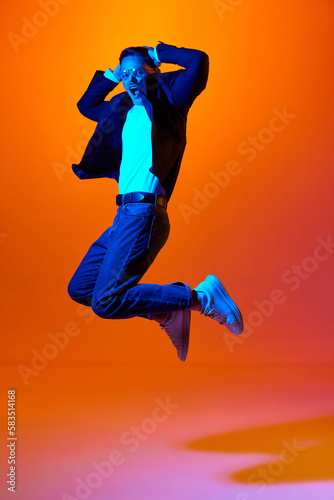 Full-length portrait of young emotive man in stylish casual clothes emotinally jumping against orange studio background in neon light.. Concept of youth, emotions, facial expression, lifestyle. Ad
