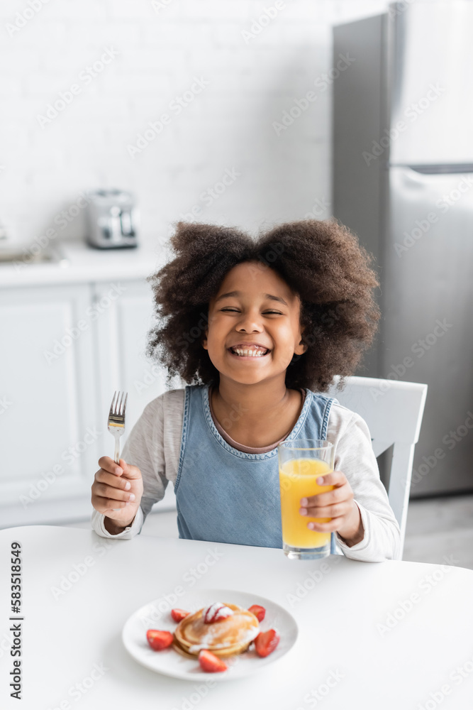 happy african american girl holding fork and glass of orange juice near pancakes while enjoying breakfast.