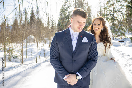 Young couple of bride and groom posing for photographer portraits in a snowy beautiful nature under high mountains High tatras on Strbske pleso with a huge hotel in behind Patria. White dress and blue © Michal