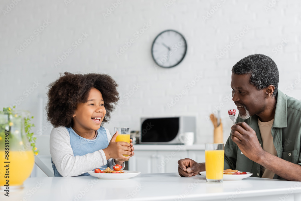 happy african american girl holding glass of orange juice while having breakfast with grandfather. happy african american girl holding glass of orange juice while having breakfast with grandfather.