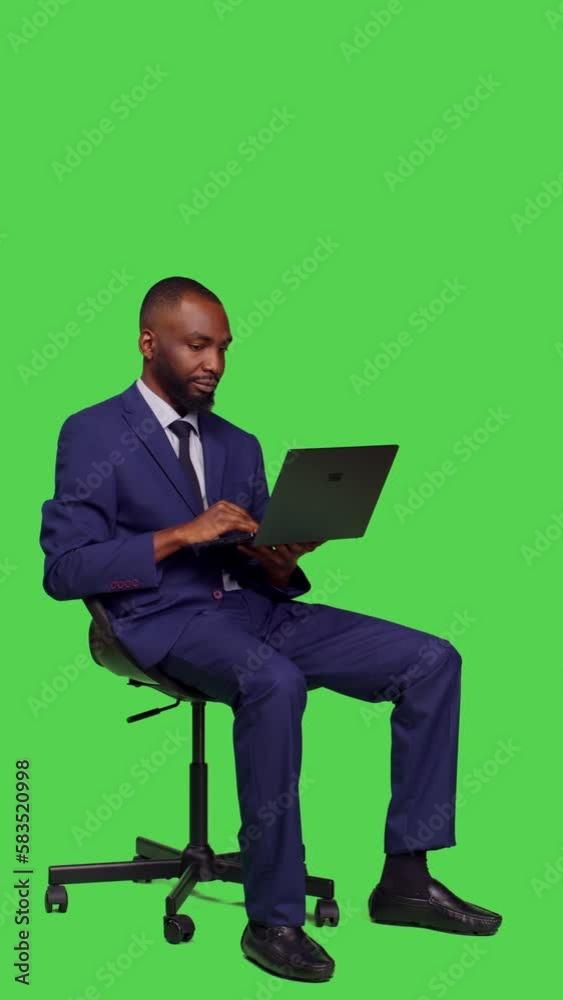 Vertical video: Side view of company worker using laptop to work on business plan, sitting on chair wearing formal suit in studio. Young manager browsing online web page on wireless computer, full Vertical video: Side view of company worker using laptop to work on business plan, sitting on chair wearing formal suit in studio. Young manager browsing online web page on wireless computer, full