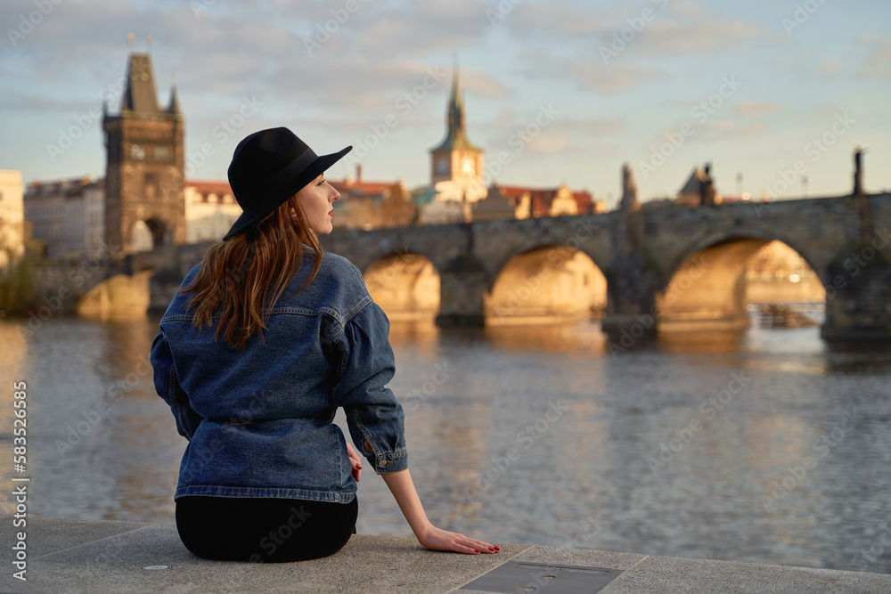 Stylish beautiful young woman wearing black hat sitting on Vltava river shore in Prague with Charles Bridge on background. Elegant retro lady fine art portrait.