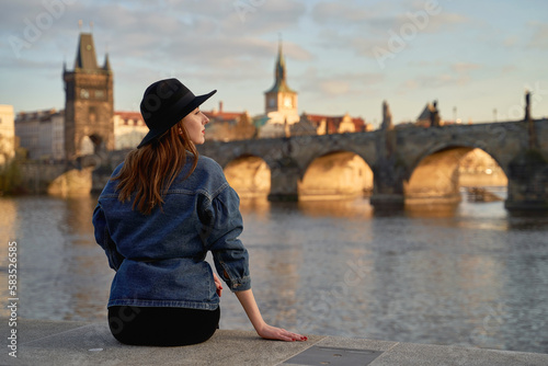 Stylish beautiful young woman wearing black hat sitting on Vltava river shore in Prague with Charles Bridge on background. Elegant retro lady fine art portrait.