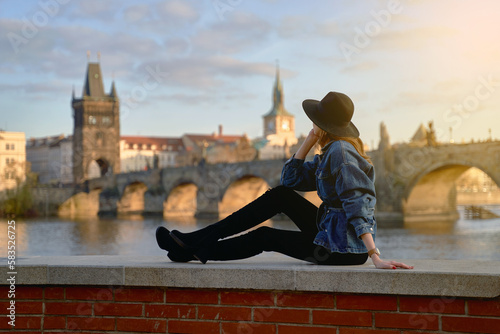 Stylish beautiful young woman wearing black hat sitting on Vltava river shore in Prague with Charles Bridge on background. Focus on foreground.