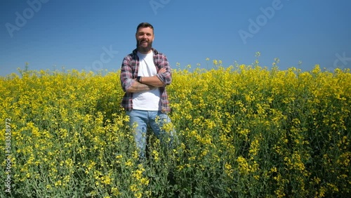 Happy and successful farmer is standing by his rapeseed field. Rapeseed plantation in bloom. Slow motion video.