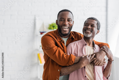 Cheerful african american man hugging mature dad and looking at camera at home. photo