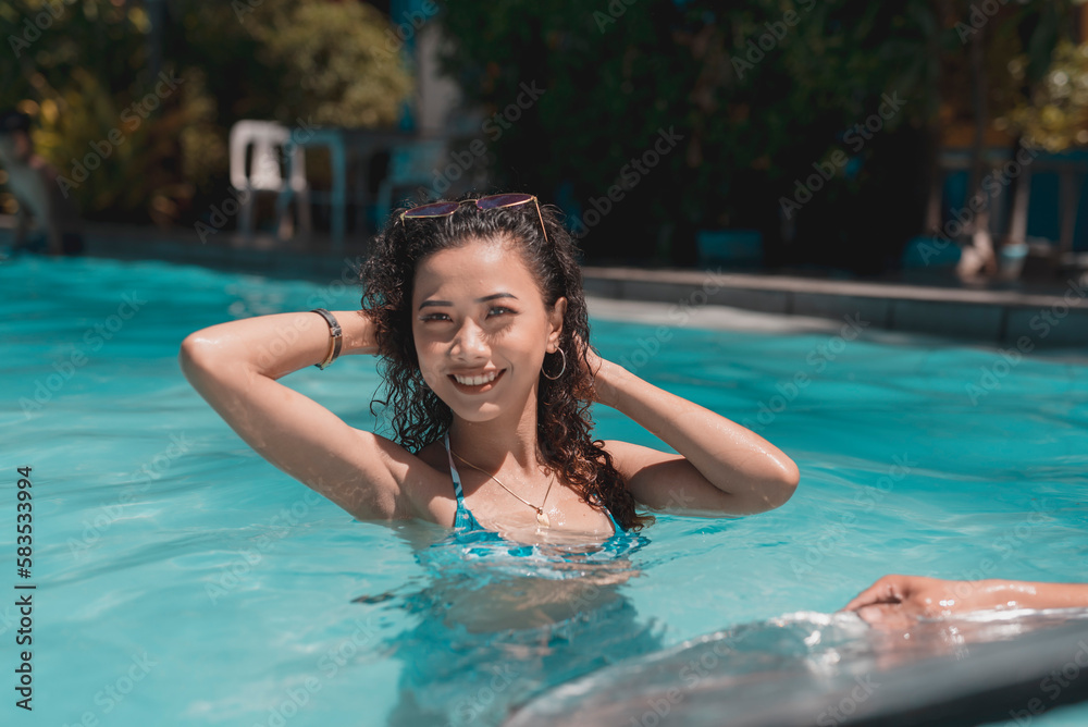 A young asian woman with curly hair relaxing in chest deep waters in the swimming pool. Enjoying her summer vacation.