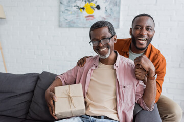 Cheerful african american middle aged man holding gift near son during father day celebration at home.