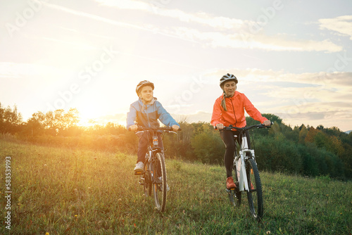Happy family outdoors, Mother and son ride a bike. Happy cute boy in helmet learn to riding a bike in park on green meadow in autumn day at sunset time.