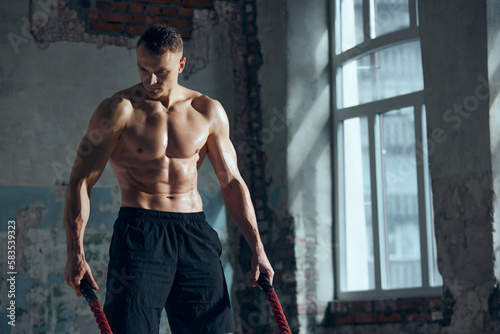 Portrait of young muscular man with strong, relief, fit body posing with sport equipments indoors. Battle ropes for workout. Concept of sportive lifestyle, body care, fitness, hobby, health, action