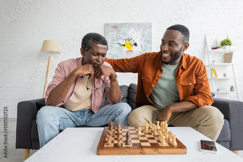 Smiling african american man hugging focused dad while playing chess in living room.