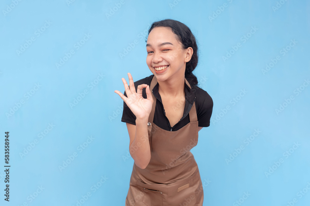 A young asian female waitress or barista winks while making the ok sign. Wearing a brown apron and black shirt welcoming a customer and against a light blue background. A young asian female waitress or barista winks while making the ok sign. Wearing a brown apron and black shirt welcoming a customer and against a light blue background.
