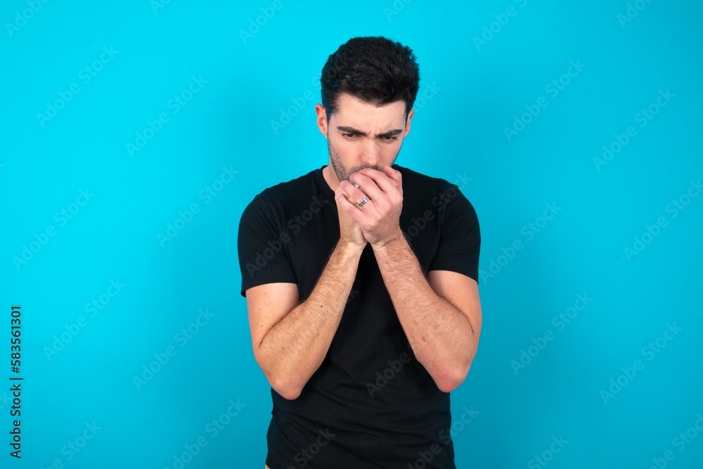 Young man wearing black T-shirt over blue studio background holding oneself, feels very cold outside, hopes that will not get cold Young man wearing black T-shirt over blue studio background holding oneself, feels very cold outside, hopes that will not get cold